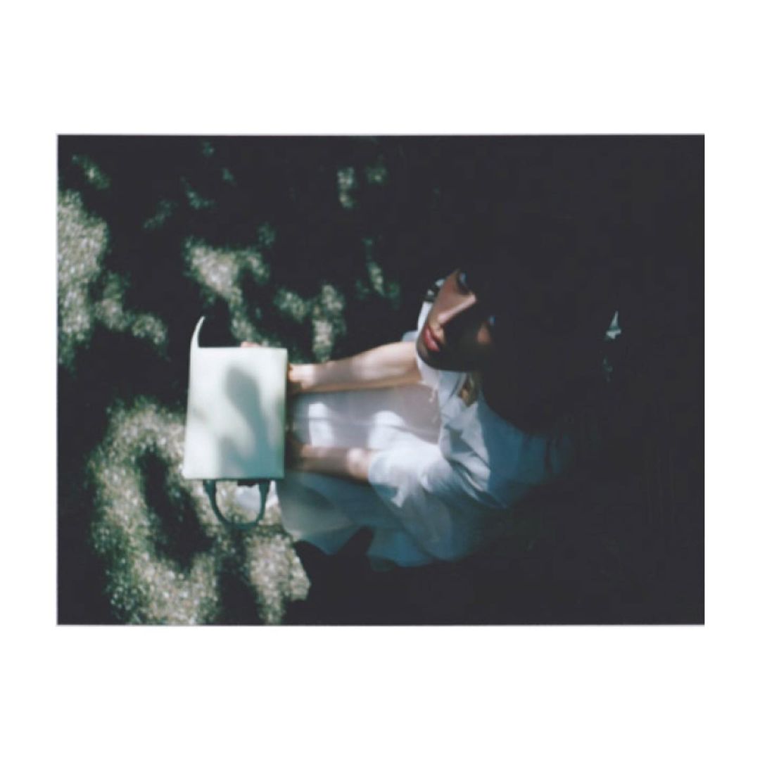 An adult woman sits outdoors with a handbag in dappled sunlight. She wears a light-colored dress. The scene is shot from overhead.