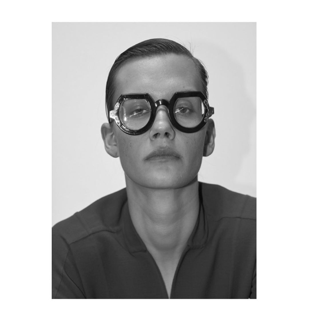 Black and white portrait of a person wearing distinctive black-framed glasses, set against a plain white backdrop in a studio setting.
