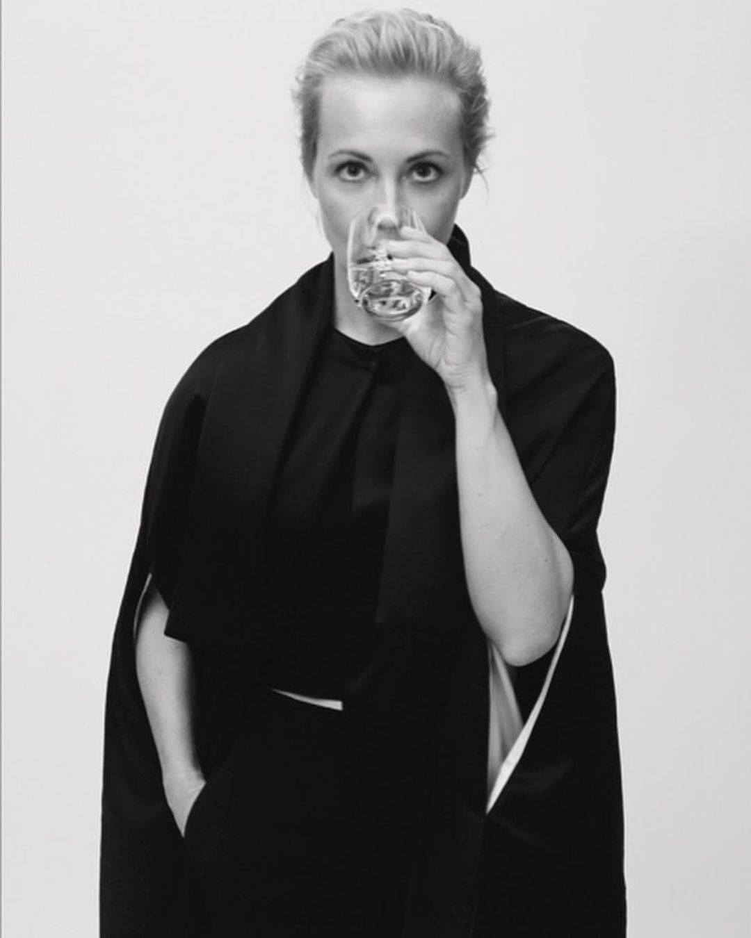 Blonde woman drinks clear liquid from glass. Dressed in dark clothing, she looks directly at the camera against a white background.