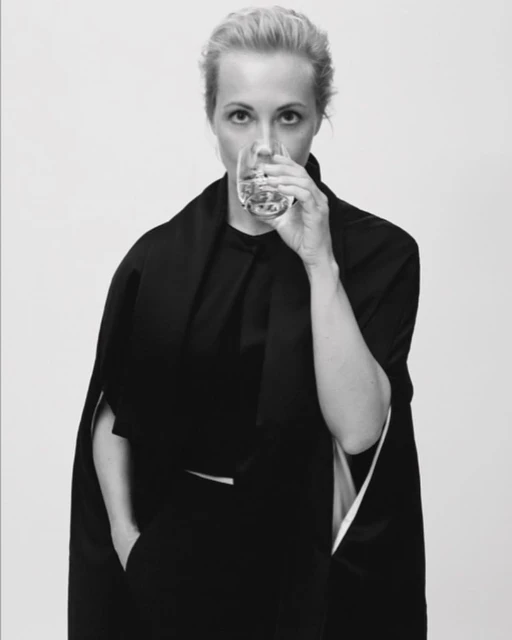 Blonde woman drinks clear liquid from glass. Dressed in dark clothing, she looks directly at the camera against a white background.