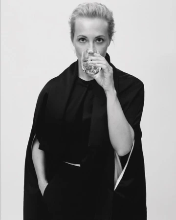 Blonde woman drinks clear liquid from glass. Dressed in dark clothing, she looks directly at the camera against a white background.