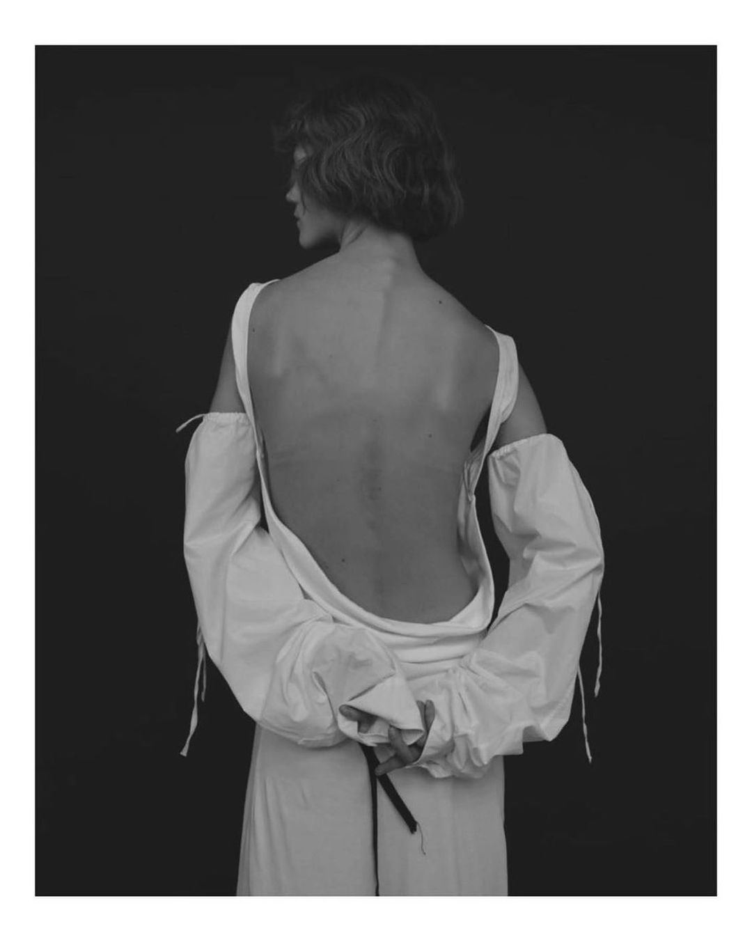 Model with short dark hair viewed from behind wearing a white open-back top against a dark background