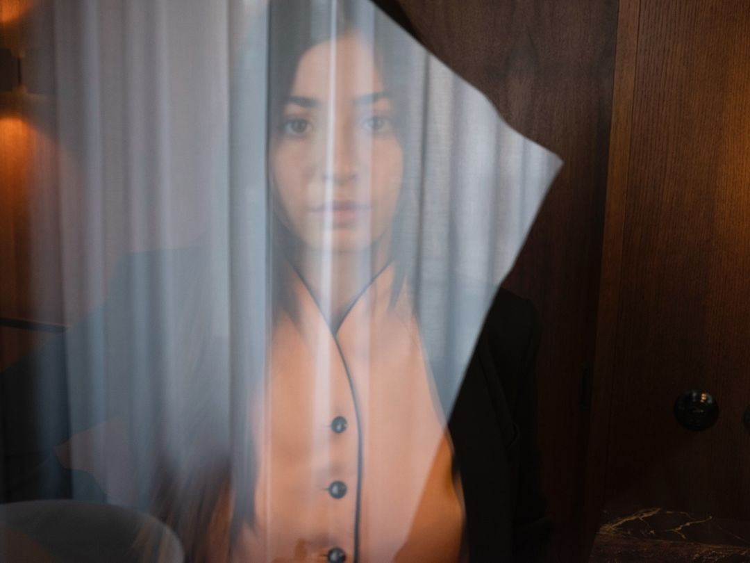 Portrait of a woman viewed through sheer vertical blinds and a tilted transparent plastic sheet in New York