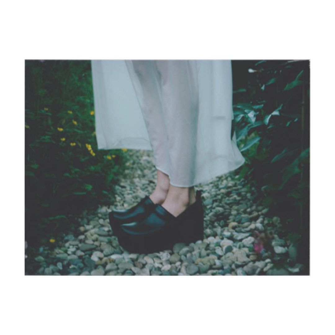 Woman in a sheer white dress and black platform shoes standing on a pebble path surrounded by greenery and small yellow flowers.