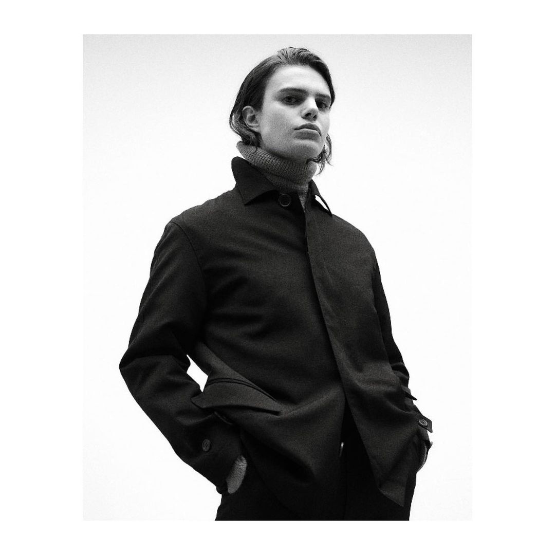 Monochrome portrait of a young adult male in a dark coat and grey turtleneck, looking directly at the camera against a white backdrop.
