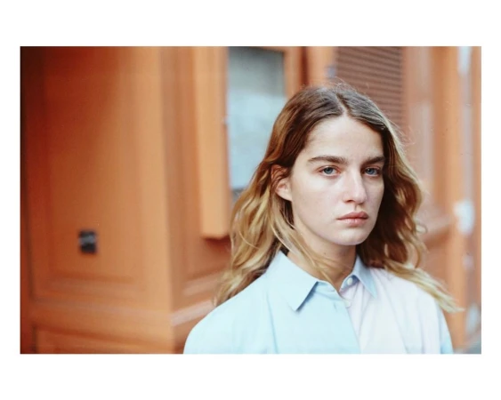 Portrait of a young woman with wavy blonde hair wearing a light blue collared shirt outdoors against a warm orange building.