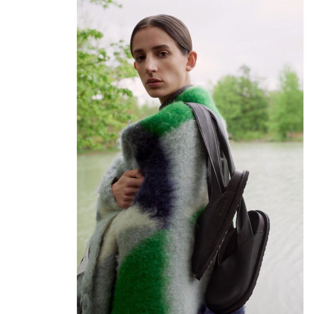 Model wearing a colourful knit coat with black slides slung over the shoulder, posing outdoors near a river.
