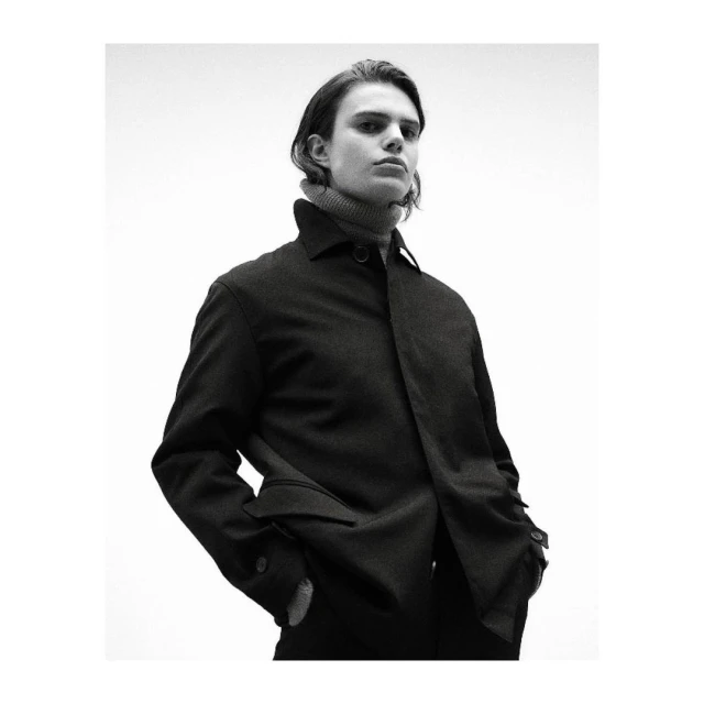 Monochrome portrait of a man wearing a dark buttoned jacket and a ribbed turtleneck against a plain background