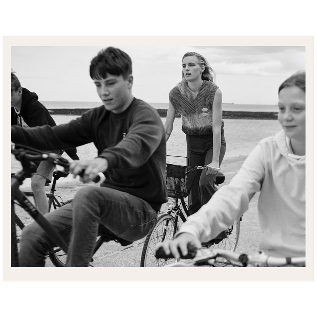 Black and white photograph of several people riding bicycles near the ocean, featuring neutral tones and a focus on outdoor activity.