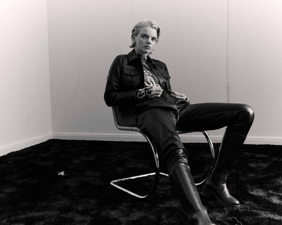 Blonde woman in black leather jacket and pants sits on a woven chair in a minimalist setting with black carpet and white walls.