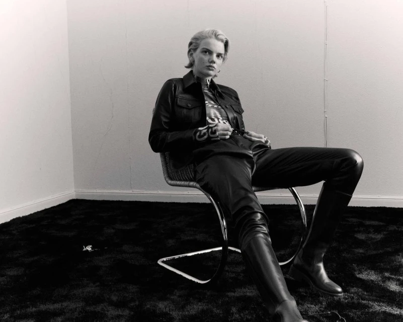 Blonde woman in black leather jacket and pants sits on a woven chair in a minimalist setting with black carpet and white walls.