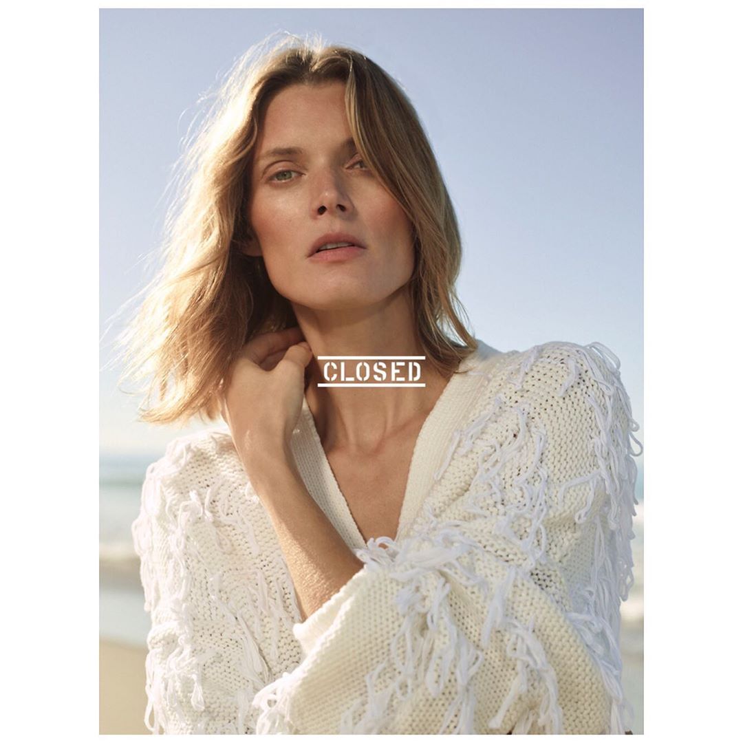 A fair-skinned woman with blonde hair poses on a beach, wearing a white, fringed knit cardigan, with the ocean and sky in the background.