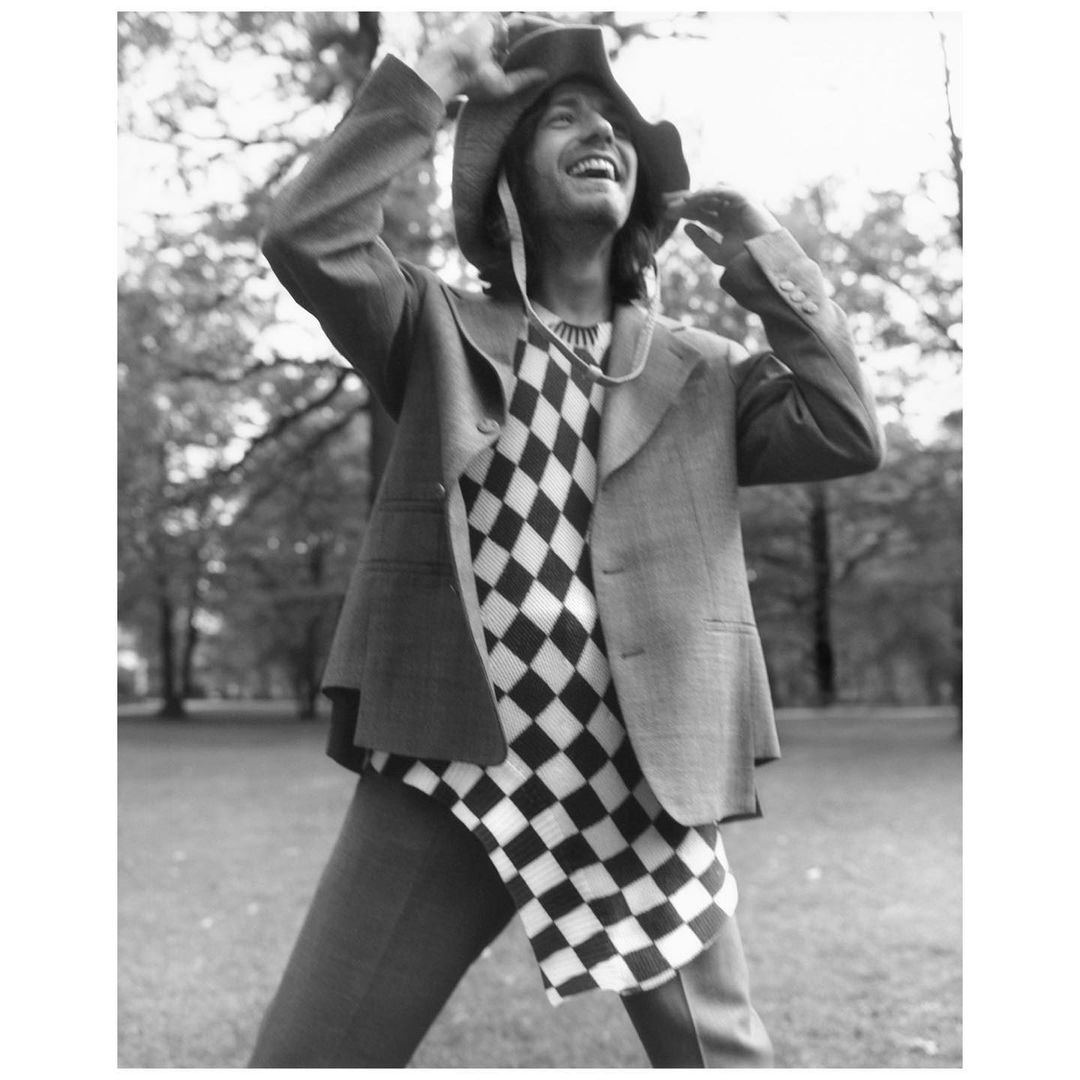 A person in a blazer, checkered shirt, and hat smiles in an outdoor park. The monochromatic photo emphasizes texture and casual fashion.
