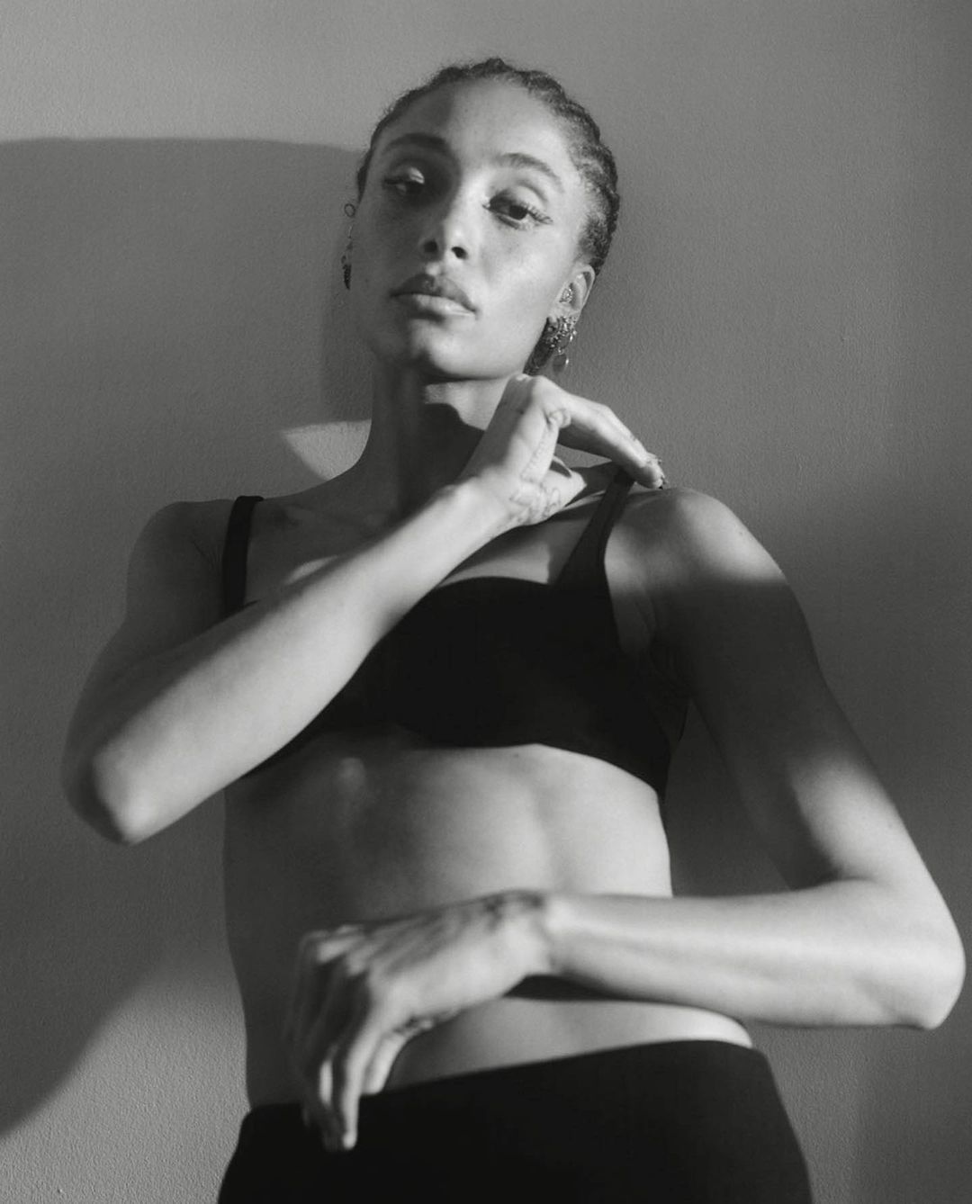 Monochrome portrait of a woman in black underwear posing against a plain wall with strong directional light and shadow.