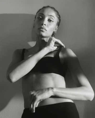 Monochrome portrait of a woman in black underwear posing against a plain wall with strong directional light and shadow.