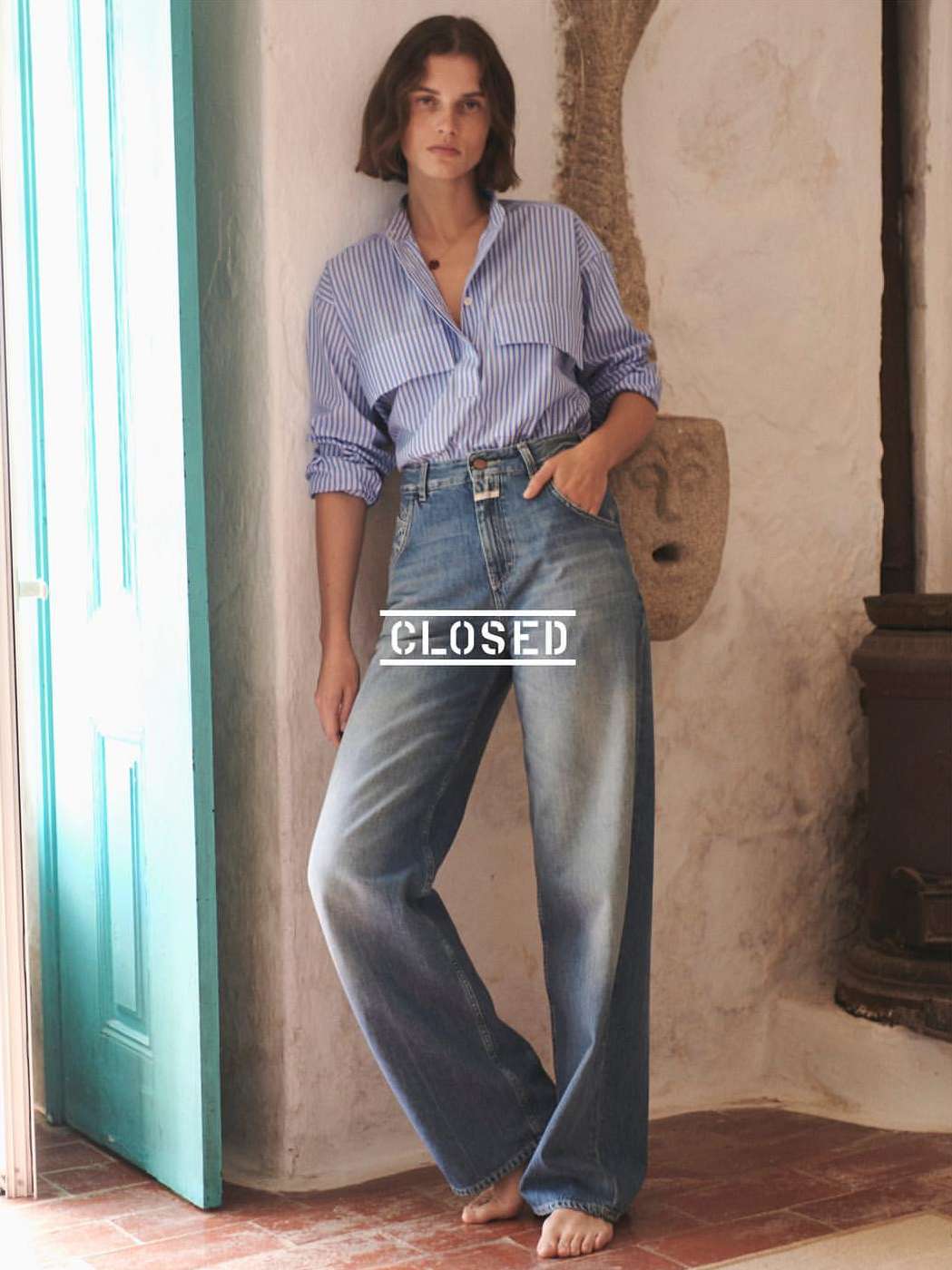 Woman in blue-striped shirt and wide-leg jeans poses indoors by turquoise door. Casual fashion shot in natural light.