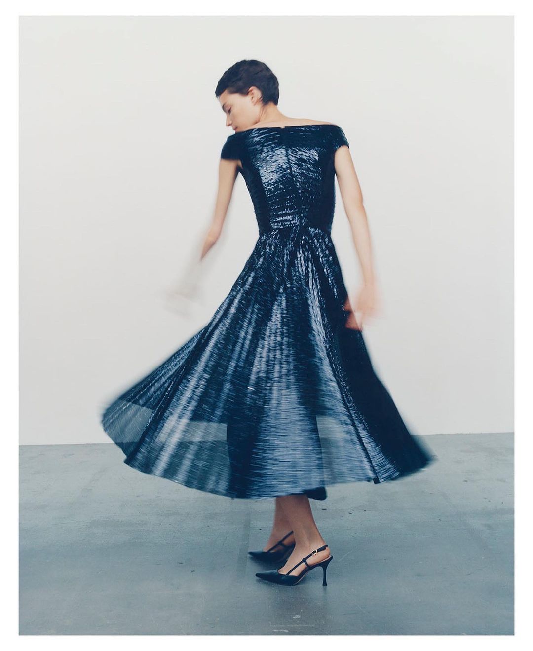 Woman in an off-the-shoulder sparkling blue midi dress and black heels is dancing in a photo studio against a white backdrop.