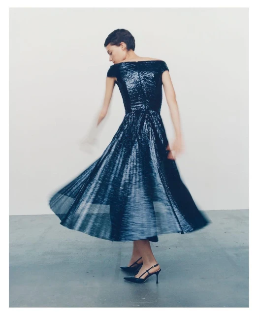 Woman in an off-the-shoulder sparkling blue midi dress and black heels is dancing in a photo studio against a white backdrop.