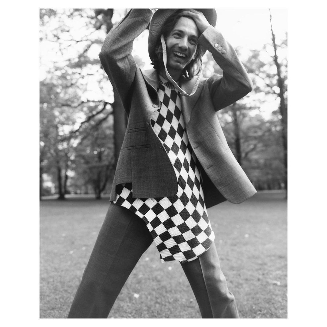 Monochrome shot of a smiling adult male outdoors in a patterned shirt, blazer, pants, and hat, standing on a grassy field with trees.