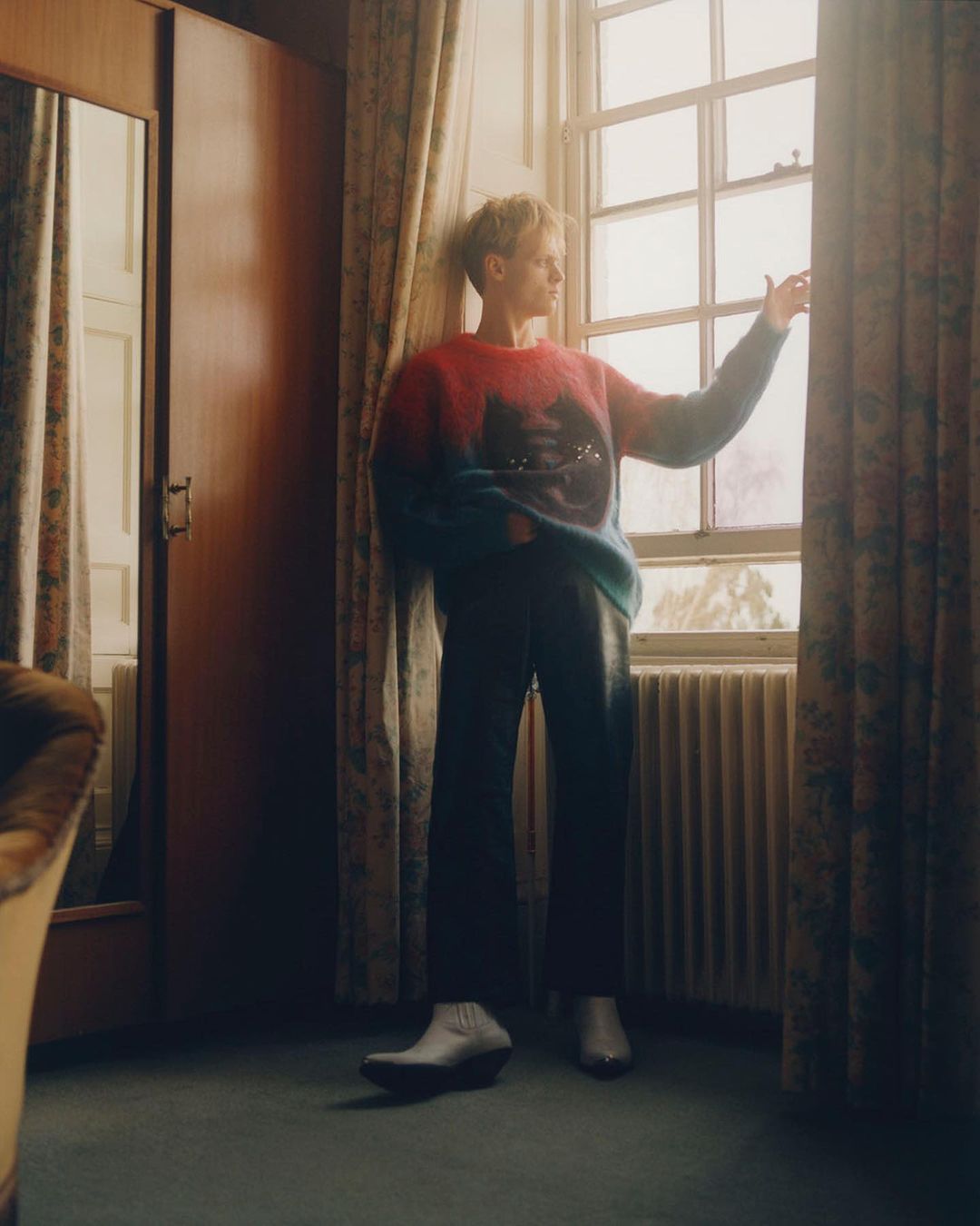 Indoor portrait of a young adult man standing near a window in a colorful sweater and dark pants, soft window light.