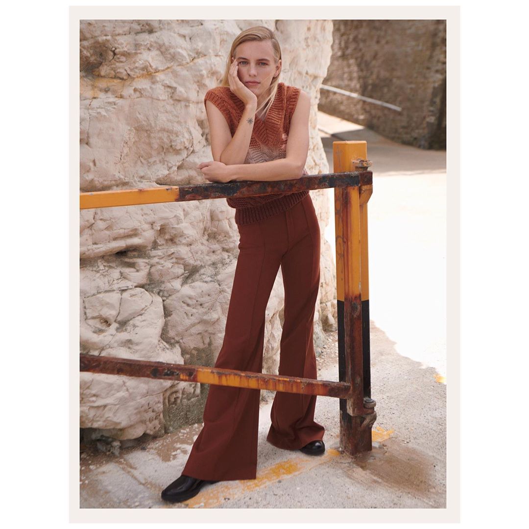 Fashion shot of a young adult female with blond hair leaning on a metal railing near a stone wall. She wears a knit top and brown trousers.