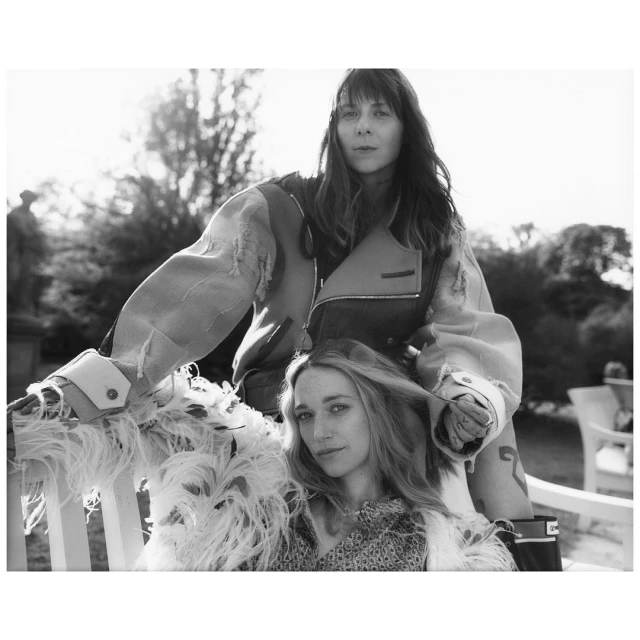 Two women in outdoor fashion shoot; one seated in ornate coat, the other standing behind in deconstructed jacket, black and white.
