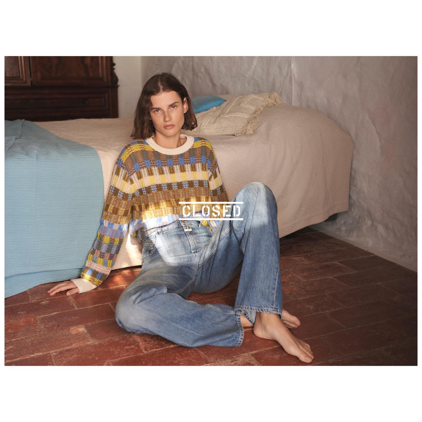 A young woman in a patterned sweater and jeans sits on a terracotta floor with a bed in the background in soft, natural light.