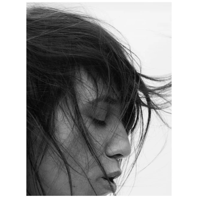 Black and white portrait of a woman with dark, windswept hair, eyes closed, and dramatic lighting creating a contrastive, artistic image.