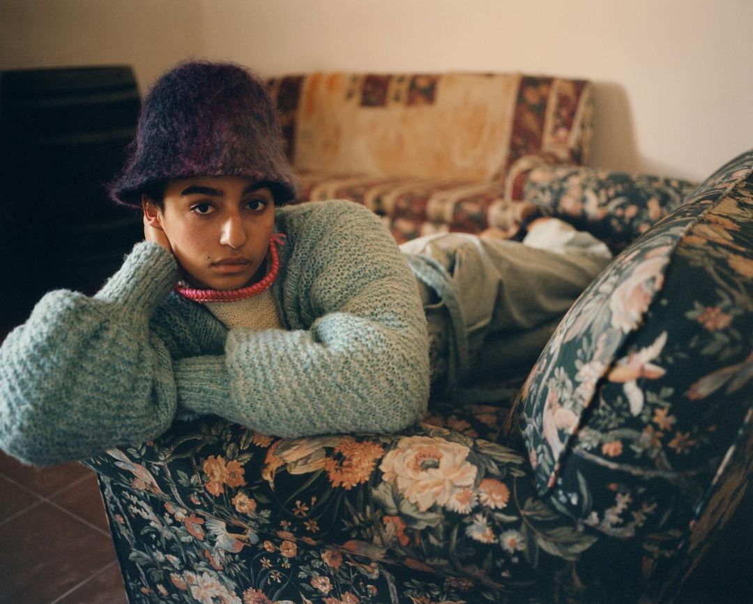 Portrait of a young person with dark hair wearing a fuzzy hat and sweater, reclining on a floral sofa, gazing intently at the camera.