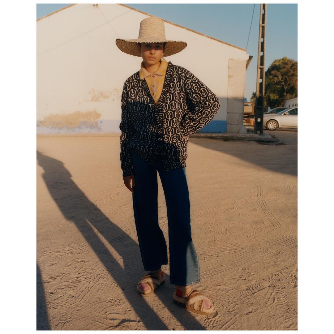 A woman poses outdoors wearing a straw sun hat, patterned cardigan, yellow collared shirt, jeans, and sandals in a sunlit, minimalist scene.