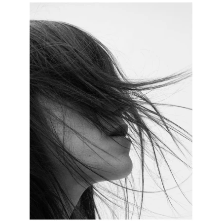 Close-up monochrome profile portrait of a woman with dark hair blowing across her face in soft light