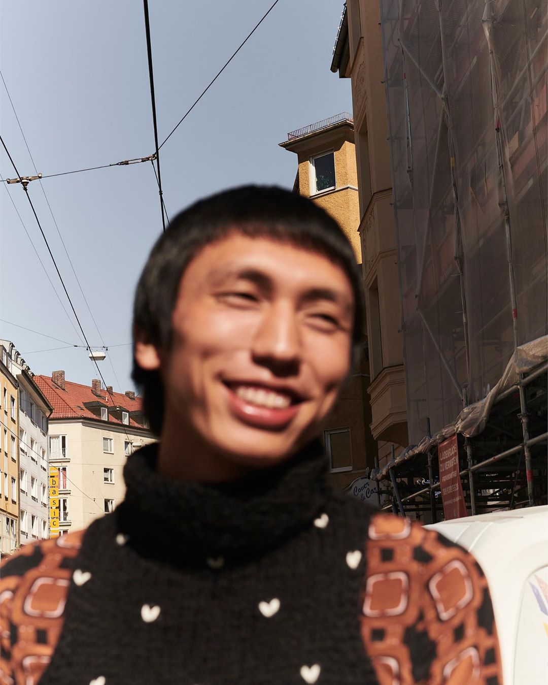 Smiling Asian man in patterned knit sweater stands on urban street with buildings in background, captured in eye-level medium shot.