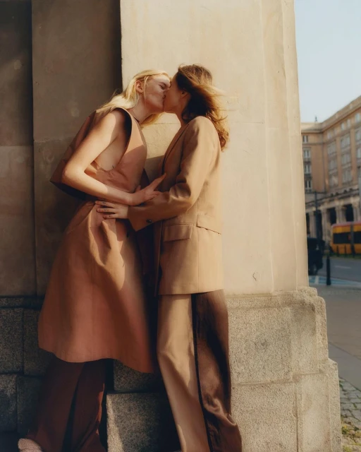 Two women in fashionable clothing embrace and kiss outdoors near a building on a sunny day, showcasing a loving relationship.
