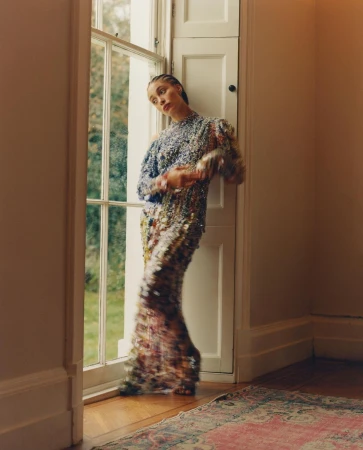 A woman in a sequined dress standing by a tall window, captured with motion blur in a sunlit room.