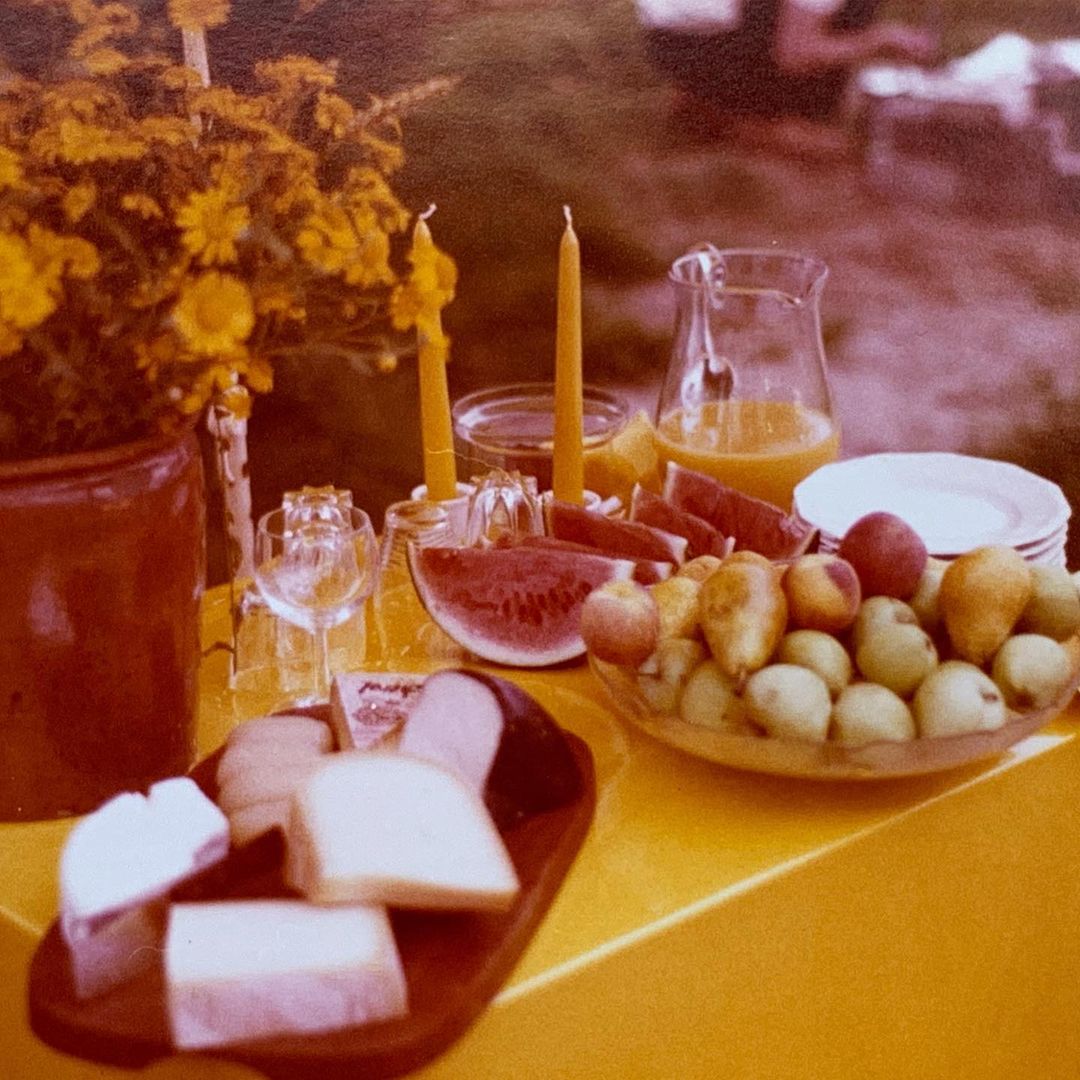 A vintage-style photograph of a yellow table laden with fruits, cheeses, and juice under natural light. Candles and flowers add to the ambiance.