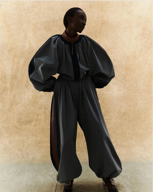 A Black woman in a grey satin oversized blouse and pants poses against a neutral background in a fashion or editorial style photograph.