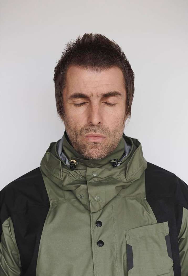 Headshot of an adult male with closed eyes, short brown hair, and wearing a green and black hooded jacket, set against a plain white background.