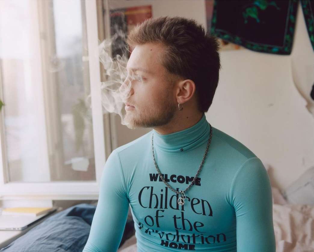 Young adult poses indoors, cigarette smoke swirls near face. Man wears a turquoise top with text. Natural window light, casual scene.