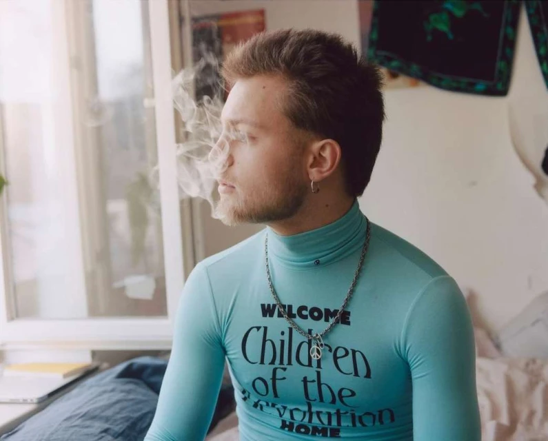Young adult poses indoors, cigarette smoke swirls near face. Man wears a turquoise top with text. Natural window light, casual scene.