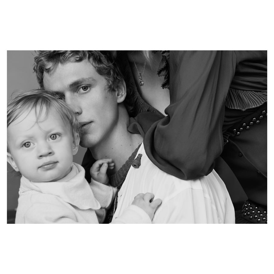 Black and white portrait of a young man holding a baby, with an adult woman partially visible behind him, creating a family-like composition.