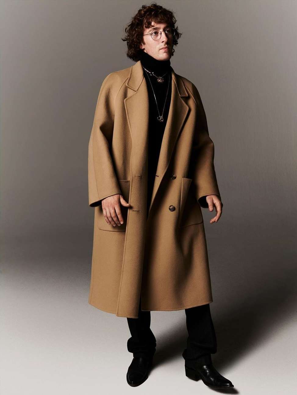 Man wearing a camel wool coat over a black turtleneck and dark trousers against a neutral studio backdrop