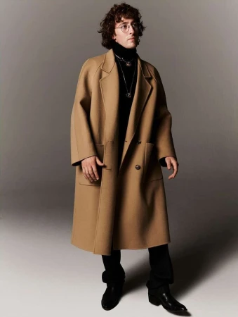 Man wearing a camel wool coat over a black turtleneck and dark trousers against a neutral studio backdrop