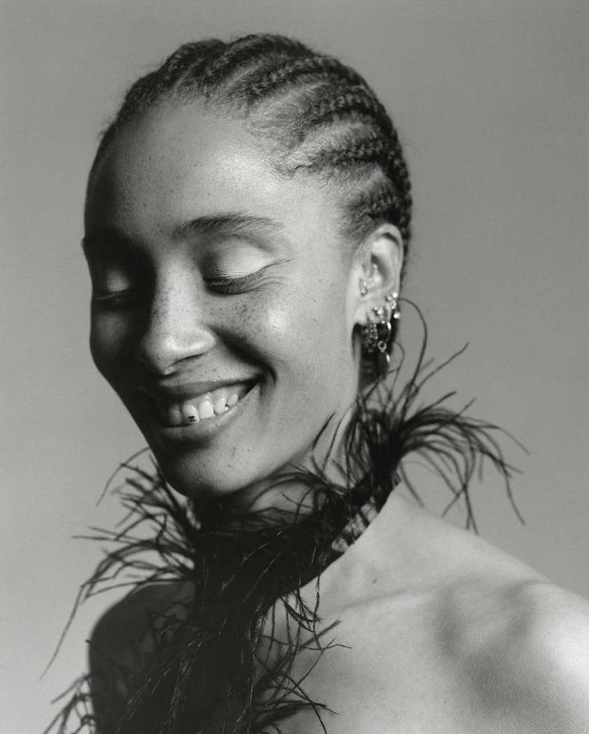 Monochrome portrait of a Black woman with braided hair, eyes closed, and a cheerful smile, accessorized with a black feather boa.