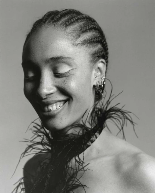 Monochrome portrait of a Black woman with braided hair, eyes closed, and a cheerful smile, accessorized with a black feather boa.