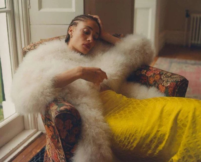 A young woman with braided hair reclines in a floral armchair wearing a fluffy white jacket and a patterned yellow dress.