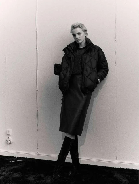 Monochrome portrait of a fair-skinned adult woman with short blond hair posing indoors wearing a black puffer jacket, black skirt, and boots.