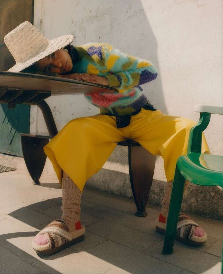 A person wearing a straw hat and colorful sweater rests on a table outdoors, with yellow pants and striped sandals adding to the casual vibe.