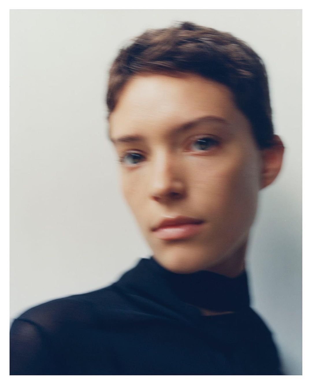 Portrait of a woman with short dark hair wearing a dark top against a plain light wall with soft focus motion blur