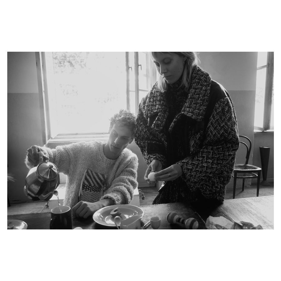 Black and white portrait of a couple at a kitchen table as one pours from a teapot, and the other peels an egg in soft lighting.