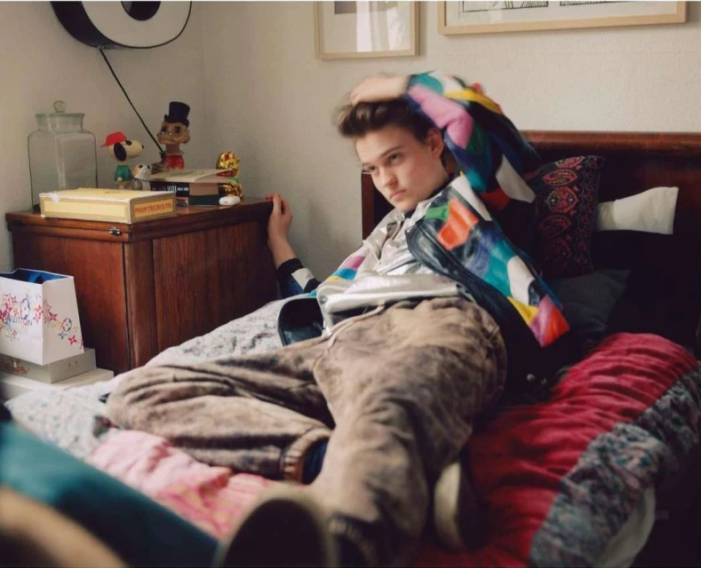 Young adult poses on bed wearing a colorful jacket and pants in a bedroom setting, lit by natural light with a casual, relaxed mood.